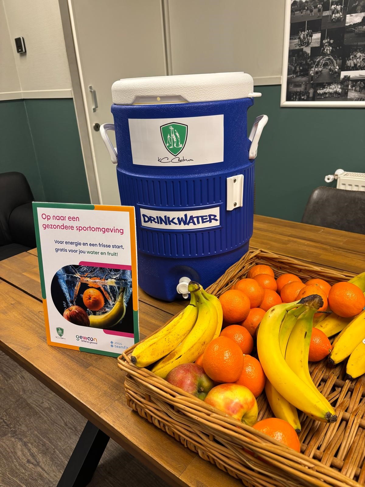 Blauwe drinkwaterton met wit deksel en handvaten en logo KC Centrum. Die staat op tafel met een schaal met verschillende soorten fruit.