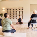 Meerdere mensen een les stoelyoga volgen onder begeleiding van een yoga instructeur