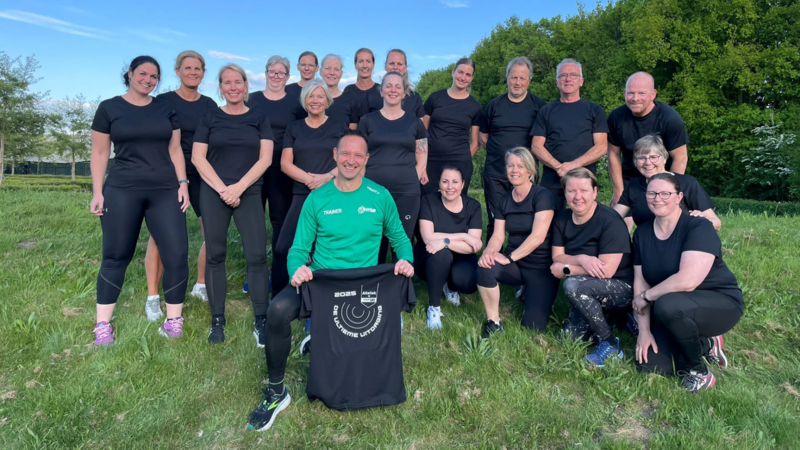 18 volwassen mensen in zwarte t-shirts en 1 volwassen man in een groene trainingsjas die poseren voor een groepsfoto