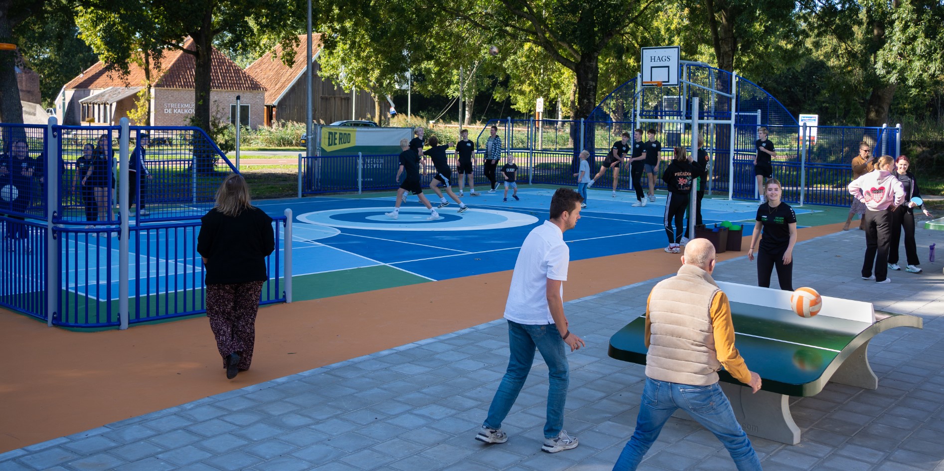 Meerdere mensen spelen en sporten op de openbare multisportvoorziening. Velden bestaan uit verschillende kleuren. Basketbalveld, voetbalveld, volleybalveld, teqbaltafel, calistenics
