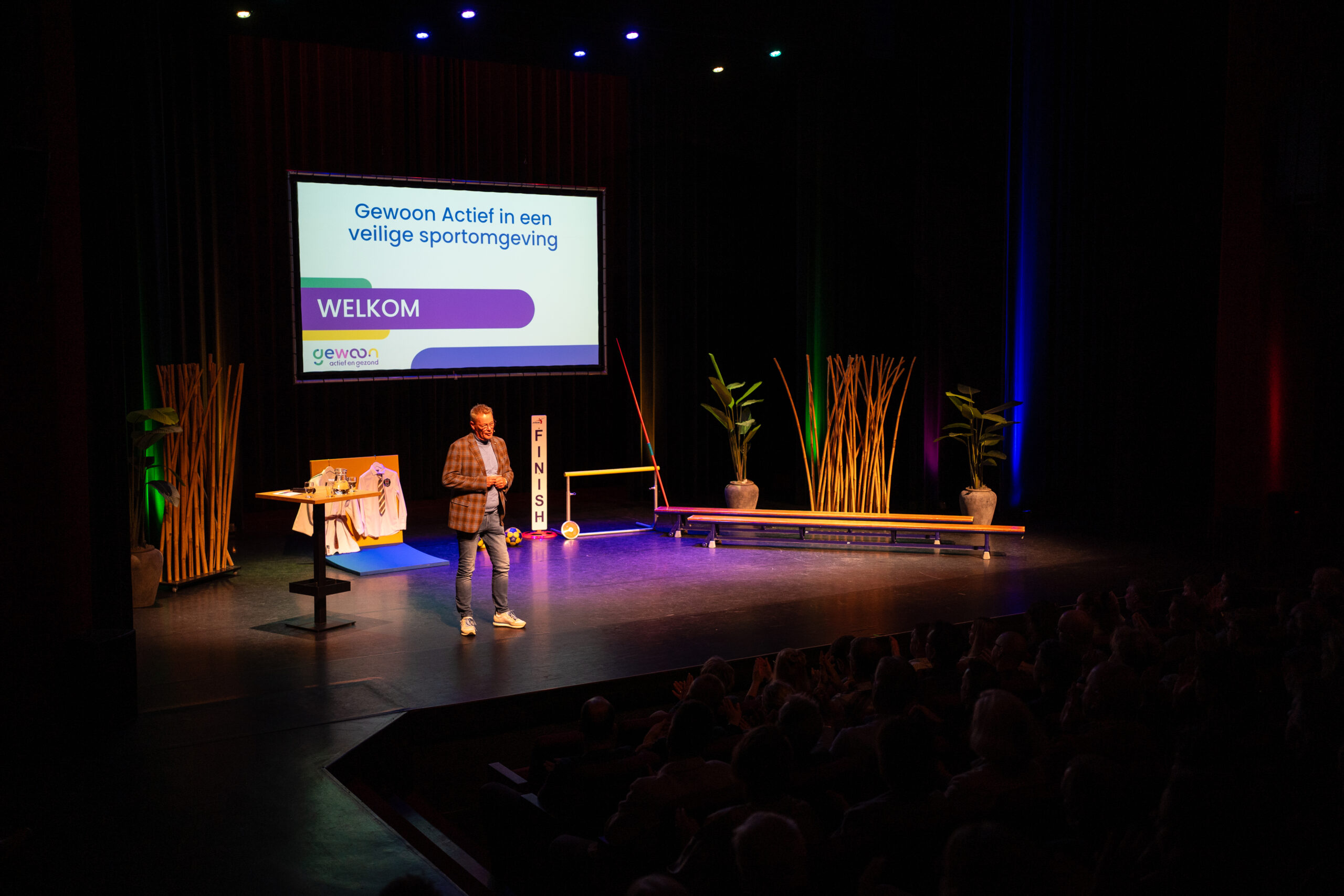 Man op theaterpodium die aangekleed is met sportattributen en gymzaalbanken en groot scherm met presentatie.