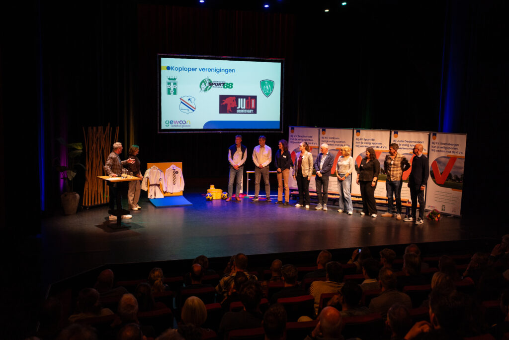 Meerdere mannen en vrouwen staan op het theaterpodium en krijgen banners uitgereikt.